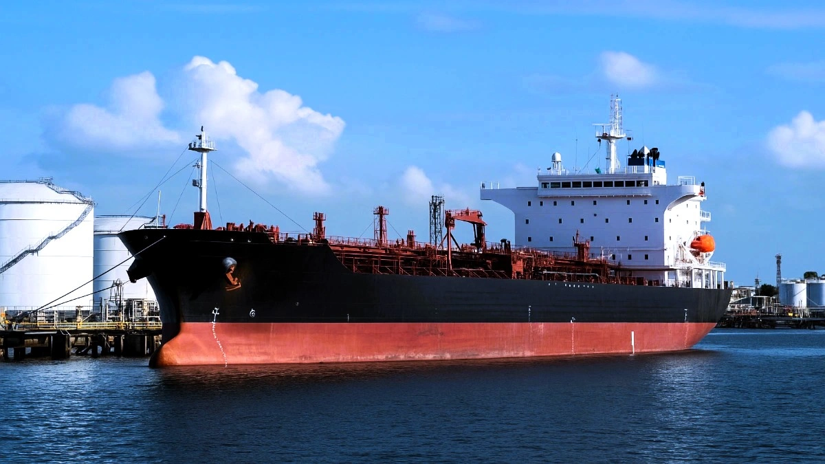 A high-resolution digital photograph of a large black and red oil tanker docked at an industrial port under clear skies. The tanker features white draft markings, red piping across the deck, and a white superstructure at the stern with navigation equipment and a bright orange lifeboat. In the background, large cylindrical fuel storage tanks and port infrastructure are visible along the shoreline.